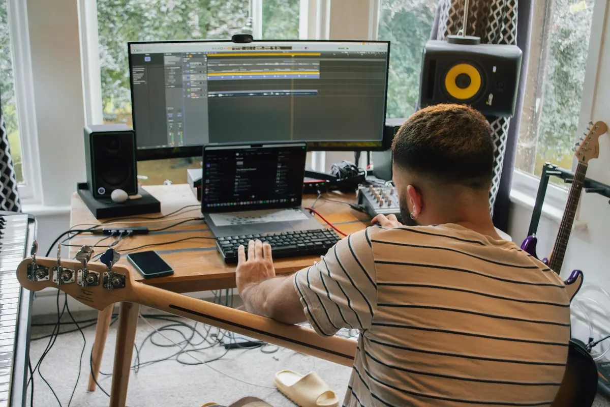 A home music studio with a desk, a big screen, speakers and guitars by the window.